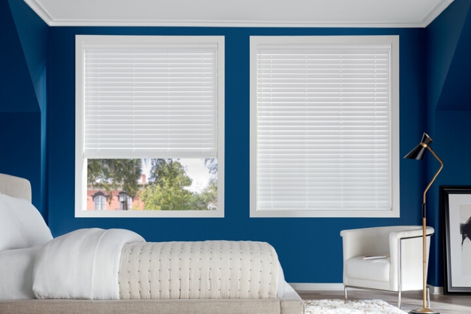 A bedroom with a bed and chair against a blue wall, featuring two white blinds covering the windows. One window shows a glimpse of trees and a building outside.